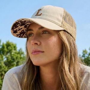 May include: A beige baseball cap featuring a brown letter "L". The underside of the peak has a cowhide print, and the back is mesh. The cap is worn by a person with long blonde hair.