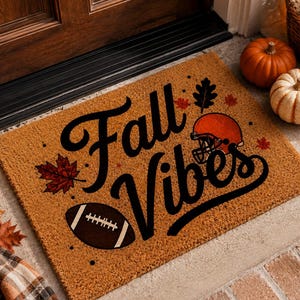 May include: A rectangular tan coir doormat featuring the black text Fall Vibes, a brown football, an orange football helmet, and several red and black maple leaf illustrations printed across the textured surface of the mat.