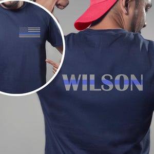 May include: Navy blue t-shirt with a gray and blue American flag graphic on the front. The back of the shirt displays the name "WILSON" in gray and blue block letters. The shirt is short-sleeved.