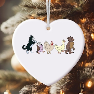 May include: A white heart-shaped Christmas ornament with a silver string. The ornament features a line of cartoon animals: a black and white cat, an owl, a chicken, a duck, and a bear. The ornament is hanging on a Christmas tree.
