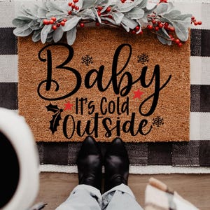 May include: A rectangular doormat with the text "Baby It's Cold Outside" in black script. The mat is brown with a decorative border and is decorated with a sprig of red berries and green leaves. Black boots and blue jeans are visible.