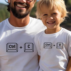 May include: Two white t-shirts with black text. One shirt reads "Ctrl + C" and the other reads "Ctrl + V". The shirts are worn by a man and a child, smiling in front of a lake and trees.