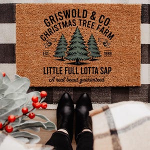 May include: A rectangular doormat with the text "Griswold & Co. Christmas Tree Farm" and "Little Full Lotta Sap" in black lettering. The design includes three green Christmas trees. The mat is brown and sits on a black and white checkered surface.