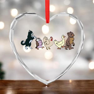 May include: Heart-shaped glass ornament with a red ribbon. The ornament features a cartoon illustration of a black and white cat, an owl, a chicken, a duck, and a bear. The background is blurred with bokeh lights.