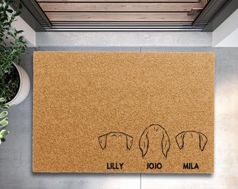 May include: A rectangular doormat made of natural brown coir fiber. The mat features three black line drawings of dog ears and the names "LILLY", "JOJO", and "MILA" below each drawing. The mat is placed in a doorway.