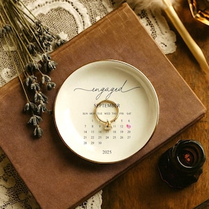 Op de afbeelding: Een wit keramisch ringschaaltje met een gouden rand, met het woord "engaged". Een gouden ring ligt op het schaaltje, dat ook een kalender voor september 2025 weergeeft. Het schaaltje ligt op een bruin boek, met gedroogde lavendel en een veer in de buurt.