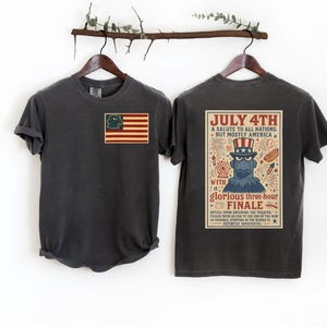 Puede incluir: Dos camisetas de color gris carb&oacute;n colgadas en un perchero de madera. Una camiseta presenta una bandera estadounidense con un personaje de dibujos animados. La otra tiene un dise&ntilde;o de p&oacute;ster vintage con el texto "July 4th" y "A salute to all nations, but mostly America."