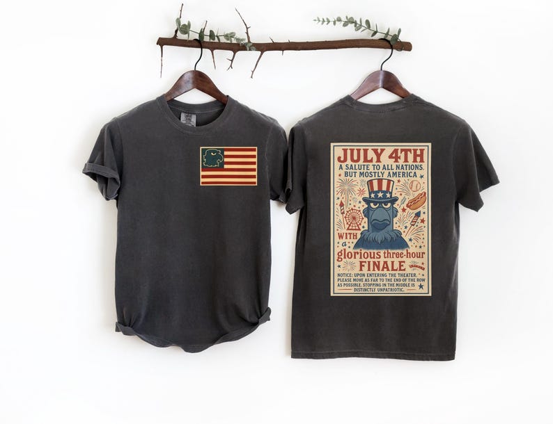 Puede incluir: Dos camisetas de color gris carb&oacute;n colgadas en un perchero de madera. Una camiseta presenta una bandera estadounidense con un personaje de dibujos animados. La otra tiene un dise&ntilde;o de p&oacute;ster vintage con el texto "July 4th" y "A salute to all nations, but mostly America."