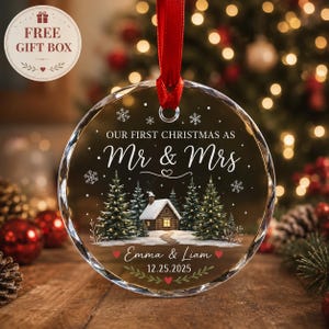 Op de afbeelding: Een ronde, doorzichtige glazen kerstornament met een rood lint. Het ontwerp toont een besneeuwd huisje met dennenbomen, sneeuwvlokken en de witte tekst "OUR FIRST CHRISTMAS AS Mr & Mrs", "Emma & Liam" en de datum "12.25.2025".