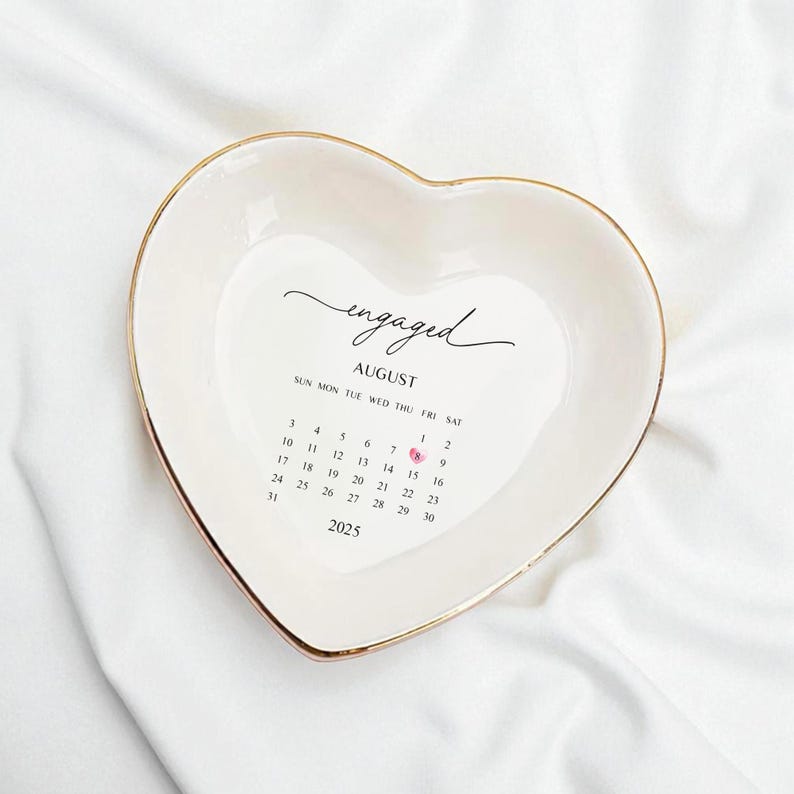 Op de afbeelding: Wit hartvormig keramisch schaaltje met een gouden rand. Het schaaltje heeft het woord "engaged" in sierlijke letters, de maand "August" en een kalender voor 2025. De 18e augustus is gemarkeerd met een roze hart.
