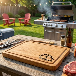 May include: A rectangular bamboo cutting board with a juice groove and the text "FATHERHOOD IS KINGDOM WORK" in black. The board is on a wooden table with grilling tools, a grill, and a bottle of beer in the background.