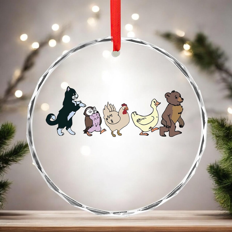May include: A clear, round ornament with a red ribbon. The ornament features a cartoon illustration of a black and white cat, owl, chicken, duck, and bear. The animals are walking in a line.