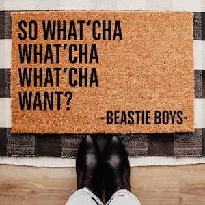 May include: A rectangular doormat made of natural coir fiber. The mat features the text "SO WHAT'CHA WHAT'CHA WHAT'CHA WANT?" in bold black letters, with "-BEASTIE BOYS-" below. The mat is placed on a black and white checkered rug.