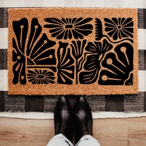 May include: A rectangular doormat with a natural brown background and a black floral design. The design features stylized flowers and leaves. The doormat is placed on a black and white checkered rug. Black boots are visible at the bottom of the image.
