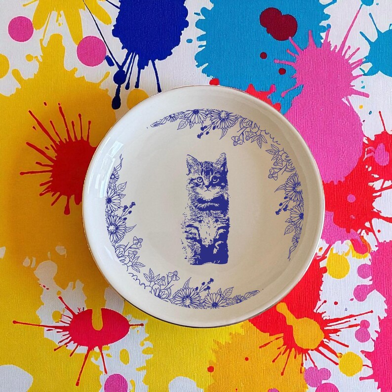 May include: A white ceramic plate with a blue illustration of a kitten and floral border. The plate has a gold rim and is set against a colorful background with paint splatters.