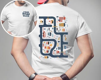 May include: White t-shirt with a navy blue graphic on the back featuring a road map design with colorful buildings and vehicles. The front of the shirt has the text "TIRED DADS CLUB" in navy blue.