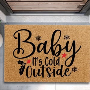 May include: A rectangular doormat with the phrase "Baby It's Cold Outside" in black script. The mat is a natural tan color, with snowflakes and red star accents. A festive welcome mat for the winter season.