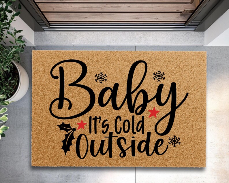 May include: A rectangular doormat with the phrase "Baby It's Cold Outside" in black script. The mat is a natural tan color, with snowflakes and red star accents. A festive welcome mat for the winter season.