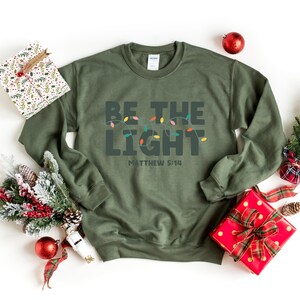 Pu&ograve; includere: Una felpa verde oliva con le parole "BE THE LIGHT" e "MATTHEW 5:14" stampate sul davanti. Il testo &egrave; sopra una fila di luci natalizie colorate. La felpa &egrave; circondata da decorazioni natalizie, tra cui regali confezionati e ornamenti.