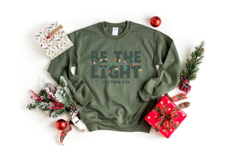 Pu&ograve; includere: Una felpa verde oliva con le parole "BE THE LIGHT" e "MATTHEW 5:14" stampate sul davanti. Il testo &egrave; sopra una fila di luci natalizie colorate. La felpa &egrave; circondata da decorazioni natalizie, tra cui regali confezionati e ornamenti.