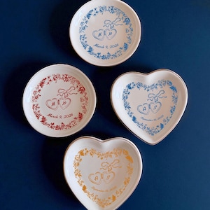 May include: Four white decorative plates with floral designs and heart-shaped details. Two plates are round, and two are heart-shaped. Each plate features initials and a date: March 9, 2026, and September 15, 2025. The plates have blue, red, and gold accents.