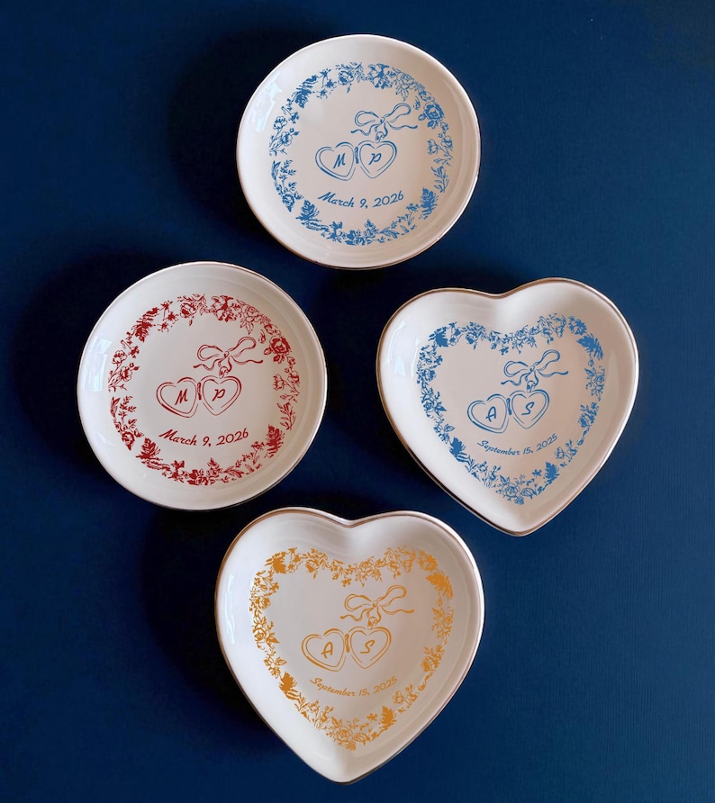 May include: Four white decorative plates with floral designs and heart-shaped details. Two plates are round, and two are heart-shaped. Each plate features initials and a date: March 9, 2026, and September 15, 2025. The plates have blue, red, and gold accents.