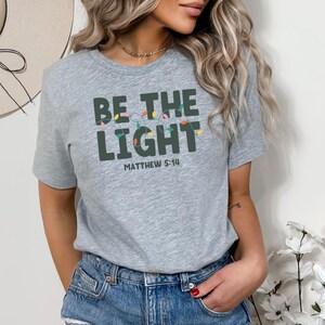 Pu&ograve; includere: T-shirt grigio chiaro con la scritta "BE THE LIGHT" in lettere verde scuro, con una fila di luci colorate sopra la parola "LIGHT" e la scritta "MATTHEW 5:14" sotto. La maglietta &egrave; abbinata a pantaloncini di jeans blu.