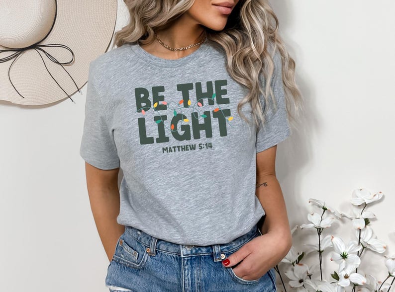 Pu&ograve; includere: T-shirt grigio chiaro con la scritta "BE THE LIGHT" in lettere verde scuro, con una fila di luci colorate sopra la parola "LIGHT" e la scritta "MATTHEW 5:14" sotto. La maglietta &egrave; abbinata a pantaloncini di jeans blu.