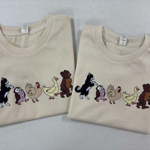 Op de afbeelding: Twee cr&egrave;mekleurige t-shirts met korte mouwen voorzien van een grafische print van een rij dieren die achter elkaar aan lopen. De reeks bestaat uit een zwart-witte kat, een kleine bruine uil, een beige kip, een gele eend en een bruine beer.
