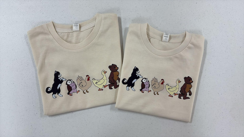 Op de afbeelding: Twee cr&egrave;mekleurige t-shirts met korte mouwen voorzien van een grafische print van een rij dieren die achter elkaar aan lopen. De reeks bestaat uit een zwart-witte kat, een kleine bruine uil, een beige kip, een gele eend en een bruine beer.