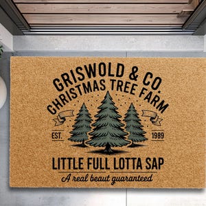 May include: A rectangular doormat with the text "GRISWOLD & CO. CHRISTMAS TREE FARM" in black, along with three green Christmas trees. The phrase "LITTLE FULL LOTTA SAP" is below the trees, with "A real beaut guaranteed" at the bottom.