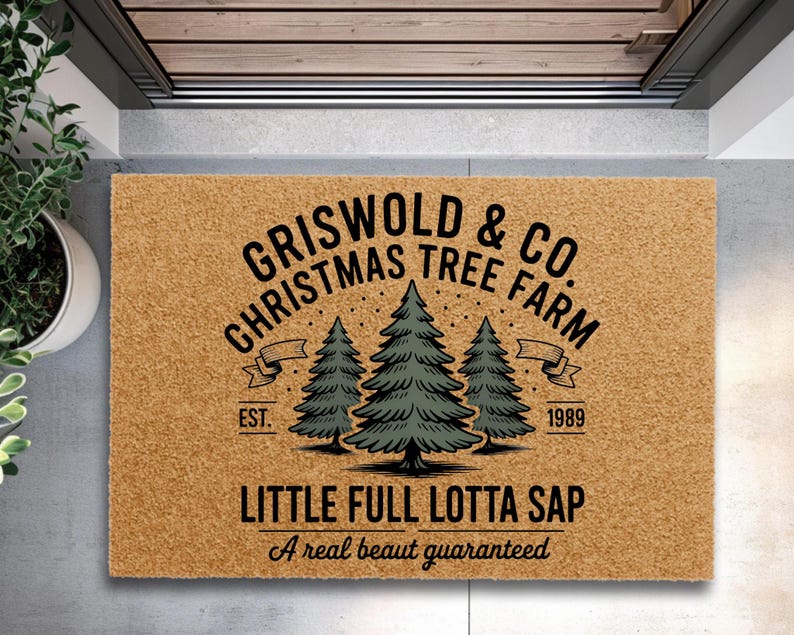 May include: A rectangular doormat with the text "GRISWOLD & CO. CHRISTMAS TREE FARM" in black, along with three green Christmas trees. The phrase "LITTLE FULL LOTTA SAP" is below the trees, with "A real beaut guaranteed" at the bottom.