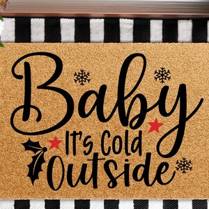 May include: A rectangular doormat with the words "Baby It's Cold Outside" in black script. The mat is a light brown color with black snowflakes, stars, and holly accents. The background has a black and white striped pattern.
