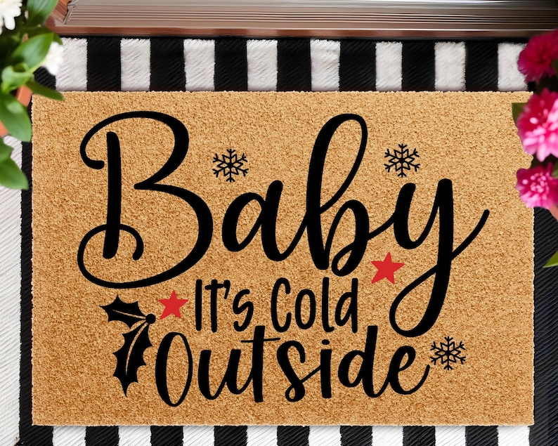 May include: A rectangular doormat with the words "Baby It's Cold Outside" in black script. The mat is a light brown color with black snowflakes, stars, and holly accents. The background has a black and white striped pattern.