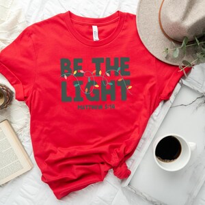 Pu&ograve; includere: Una maglietta rossa con la scritta "BE THE LIGHT" in verde scuro, con una fila di luci colorate. Il testo "Matthew 5:16" &egrave; sotto. Un cappello dai toni neutri, un vassoio con una tazza di caff&egrave; e un libro sono anche nell'immagine.