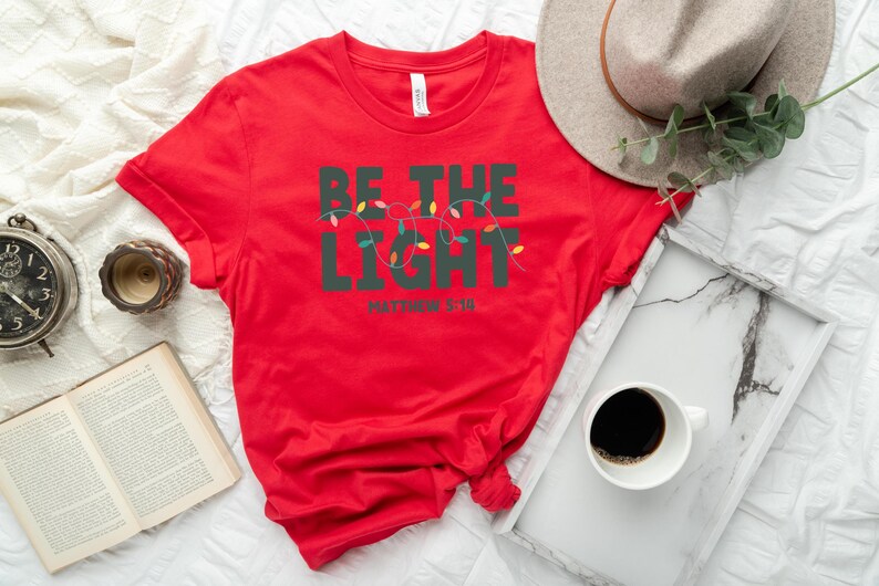 Pu&ograve; includere: Una maglietta rossa con la scritta "BE THE LIGHT" in verde scuro, con una fila di luci colorate. Il testo "Matthew 5:16" &egrave; sotto. Un cappello dai toni neutri, un vassoio con una tazza di caff&egrave; e un libro sono anche nell'immagine.
