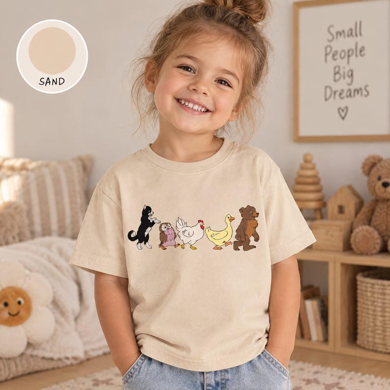 Op de afbeelding: Een zandkleurig t-shirt met een grafische print van een zwart-witte kat, een kleine uil, een witte kip, een gele eend en een bruine beer die in een rij lopen. Het t-shirt heeft een ronde halslijn en korte mouwen, met een cirkelvormig label.