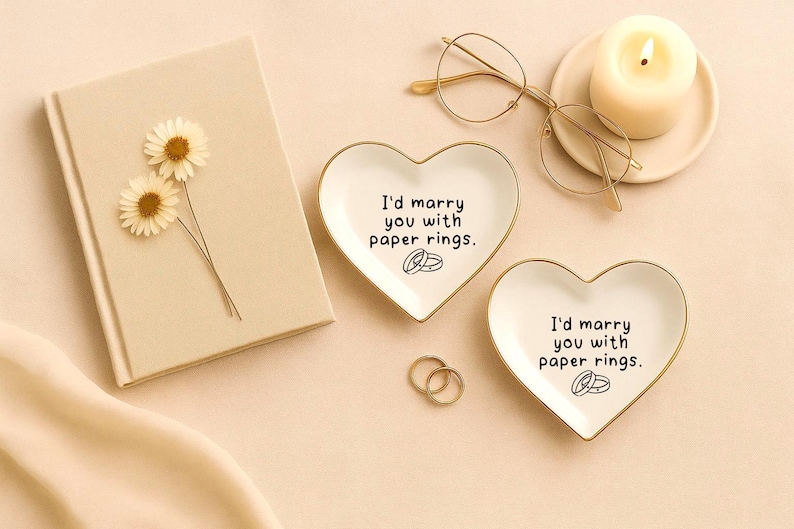 May include: A romantic still life featuring heart-shaped ring dishes with the text "I'd marry you with paper rings." Also includes a beige book with dried flowers, gold-rimmed glasses, a lit candle, and gold rings.