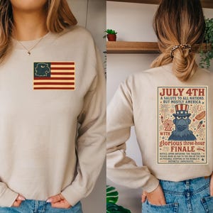 Puede incluir: Sudadera beige con un dise&ntilde;o patri&oacute;tico. La parte delantera presenta una bandera estadounidense con una cabeza de &aacute;guila en el campo azul. La parte trasera muestra un p&oacute;ster de estilo vintage con el texto "July 4th" y un &aacute;guila de dibujos animados.