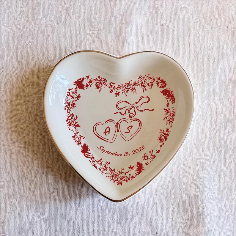 May include: A heart-shaped ceramic dish with a gold rim. The white dish has a red floral design around the edge and two connected hearts with the initials "A" and "S". The date "September 15, 2025" is printed on the dish.