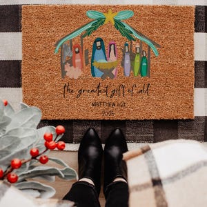 May include: A rectangular doormat with a Nativity scene illustration. The image features a stable with figures in blue, pink, and green, with the text "the greatest gift of all" and "Matthew 1:21 2025". The mat is brown.