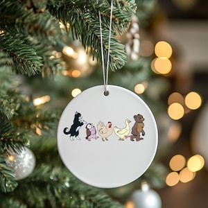 May include: A white ceramic ornament with a silver string for hanging. The ornament features a cartoon illustration of a black cat, owl, chicken, duck, and bear. The ornament is hanging on a Christmas tree.