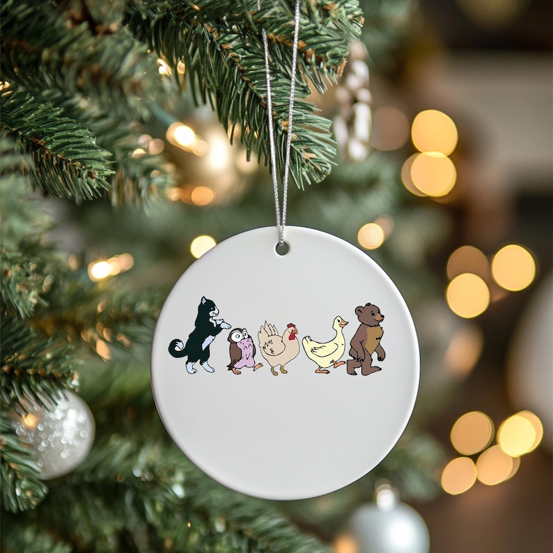 May include: A white ceramic ornament with a silver string for hanging. The ornament features a cartoon illustration of a black cat, owl, chicken, duck, and bear. The ornament is hanging on a Christmas tree.