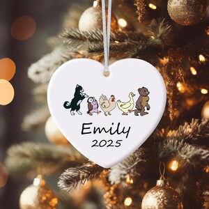 May include: White heart-shaped Christmas ornament with a silver string. The ornament features cartoon illustrations of a cat, owl, chicken, duck, and bear. The name "Emily" and the year "2025" are printed in black.