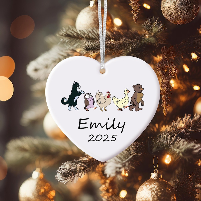 May include: White heart-shaped Christmas ornament with a silver string. The ornament features cartoon illustrations of a cat, owl, chicken, duck, and bear. The name "Emily" and the year "2025" are printed in black.