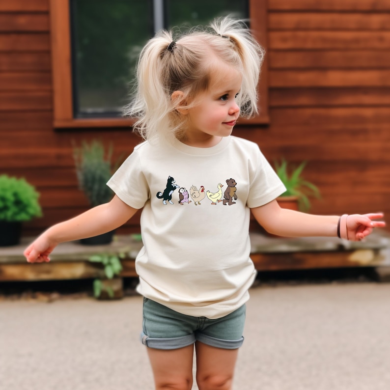 Op de afbeelding: Een cr&egrave;mekleurig T-shirt met korte mouwen en een horizontale grafische print van vijf kleine dieren op een rij, waaronder een zwart-witte kat, een kleine uil, een kip, een gele eend en een bruine beer, gecombineerd met een grijze spijkerbroek.