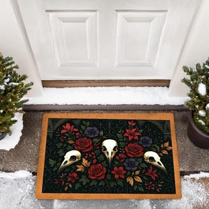 May include: A rectangular doormat with a dark background, featuring a floral design with red roses, purple flowers, and three bird skulls. The mat has a brown border and is placed in front of a white door.