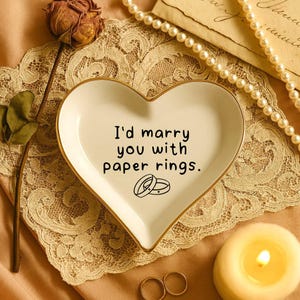 May include: A heart-shaped, white ceramic trinket dish with gold trim, featuring the text "I'd marry you with paper rings." and two ring illustrations. The dish rests on a lace doily, with a pearl necklace and a lit candle nearby.