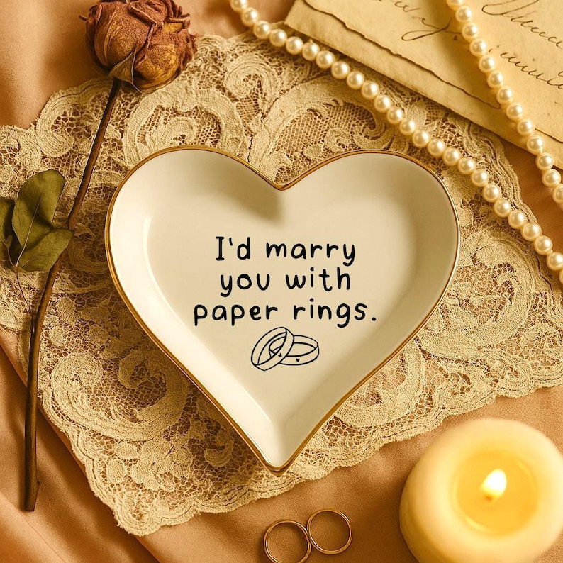 May include: A heart-shaped, white ceramic trinket dish with gold trim, featuring the text "I'd marry you with paper rings." and two ring illustrations. The dish rests on a lace doily, with a pearl necklace and a lit candle nearby.