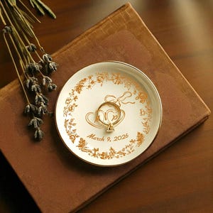 May include: A white ceramic ring dish with gold floral and heart designs, holding a gold ring with a diamond. The dish has a gold rim and the inscription "March 9, 2026". The dish rests on a brown book.